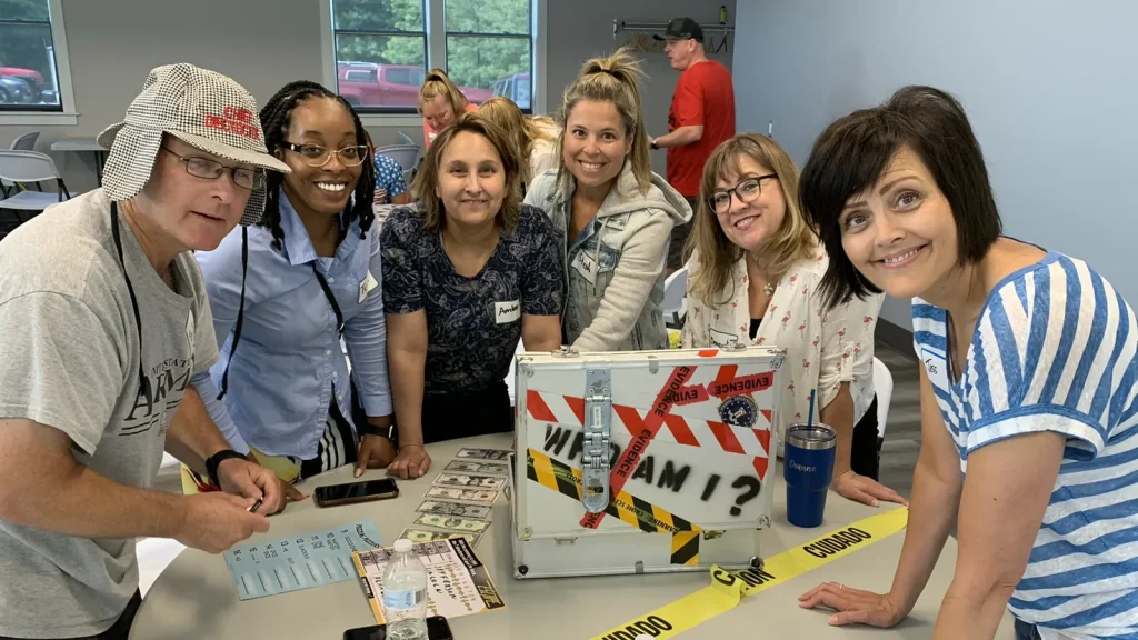 Six adults gather around a table, smiling at a silver mystery box taped with red-and-white caution stripes reading 'Who Am I?'