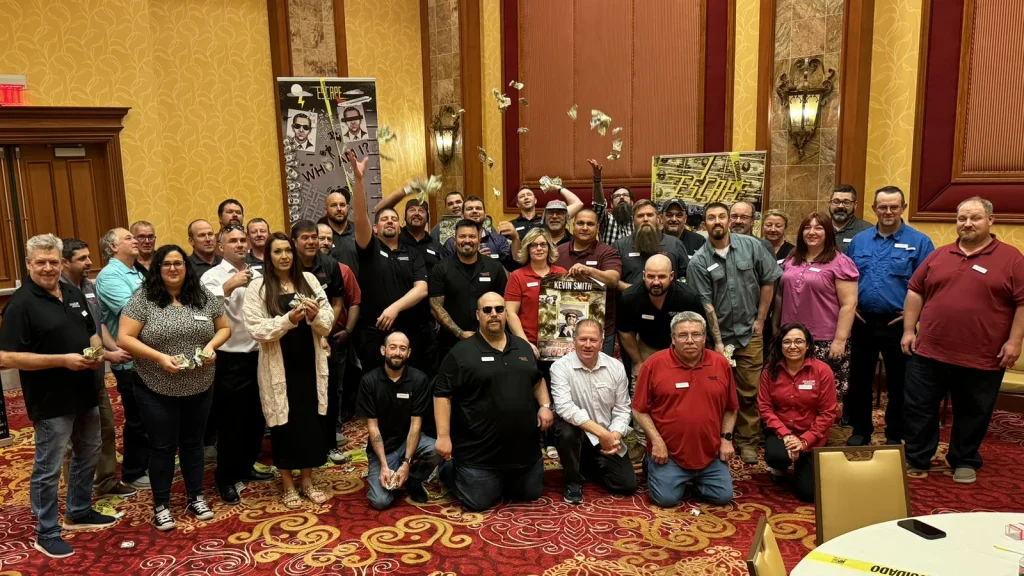 Large group of people in a banquet hall posing for a group photo, some holding cash and throwing bills in the air, festive posters in the background.