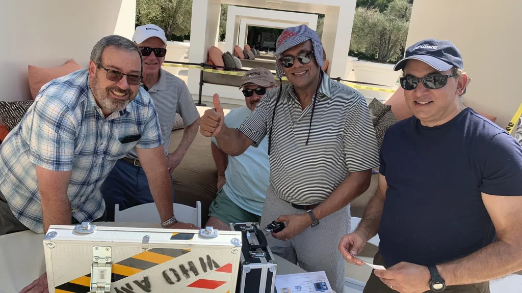Five men outdoors around a metal equipment case, smiling at the camera; one gives a thumbs-up gesture.