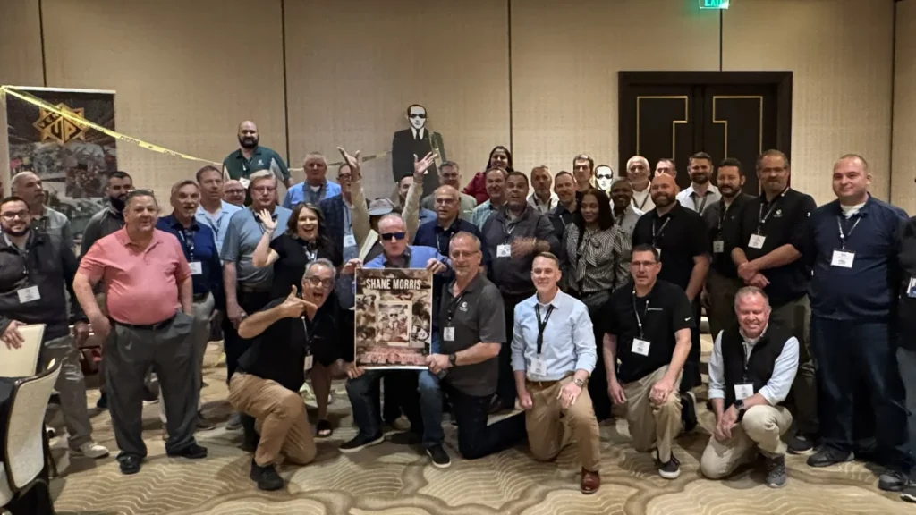 Group of conference attendees posing for a group photo; one person holds a framed poster while caution tape decorates the backdrop in a banquet hall.
