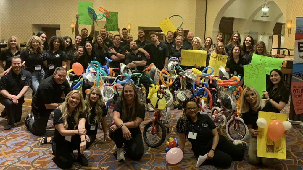 Group of coworkers posing with colorful DIY bicycle sculptures and signs at an indoors event, smiling at the camera.