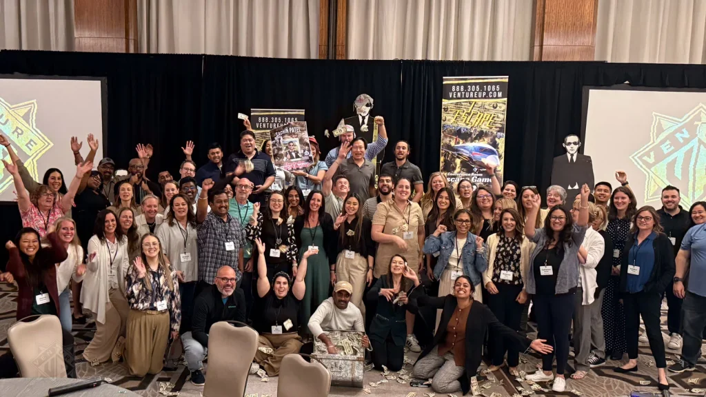 Large group of people posing for a photo at a conference, many raising hands, with banners on stage behind them.
