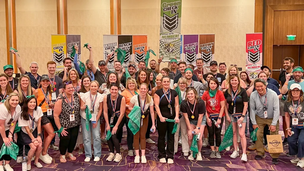 Large group photo of attendees at a conference, many wearing medals and green bandanas, posing in a banquet hall with colorful 'Check In' banners in the background.