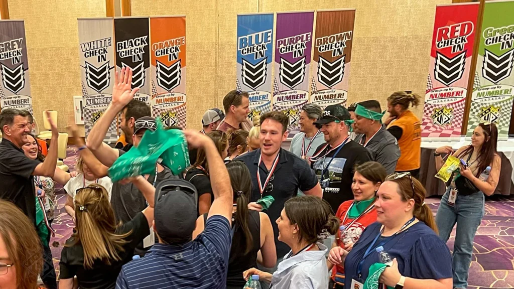 Group of attendees celebrating in a conference hall near colorful Check‑In banners, some wearing medals and high‑fiving.