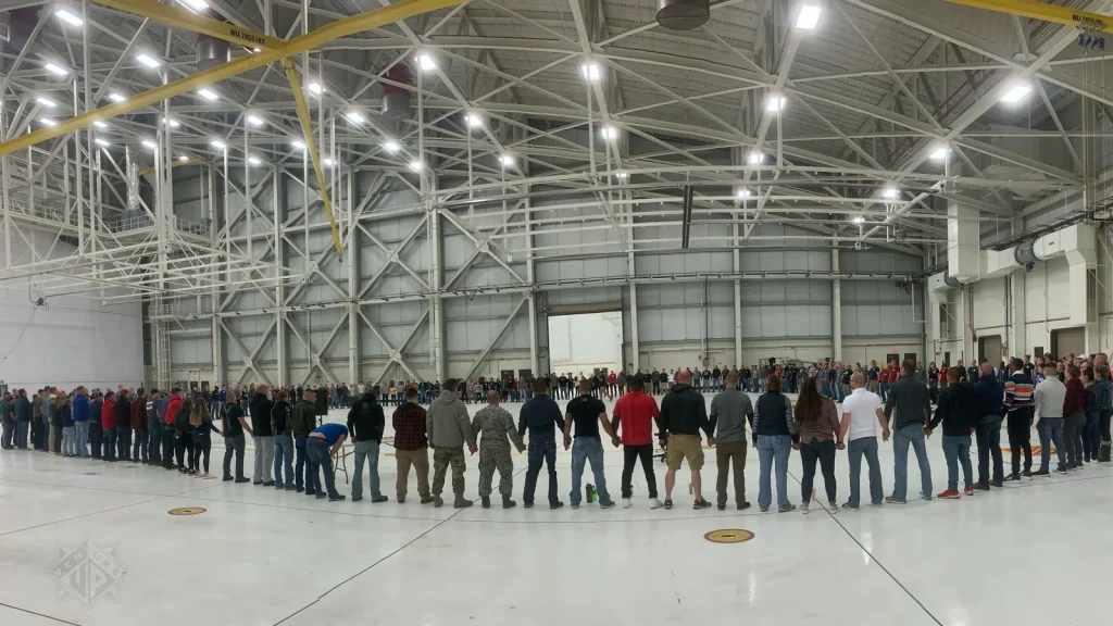 People form a large circle in a spacious aircraft hangar, holding hands around the edge.