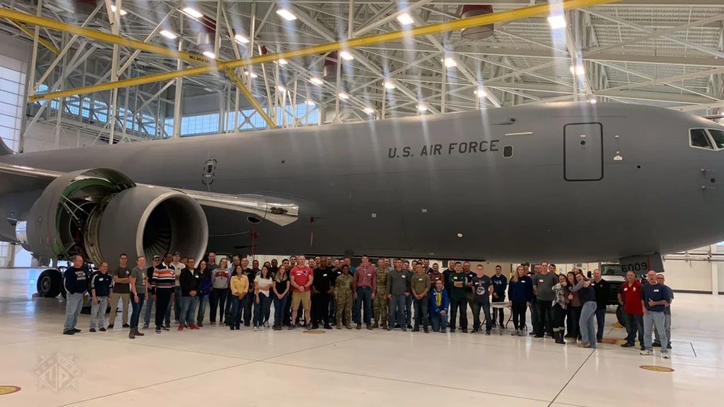 Group of people posing in front of a large gray U.S. Air Force aircraft inside a hangar, with an exposed engine nacelle visible to the left.