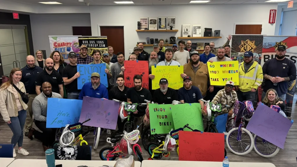 A large group poses behind colorful child-size bicycles and handmade signs.