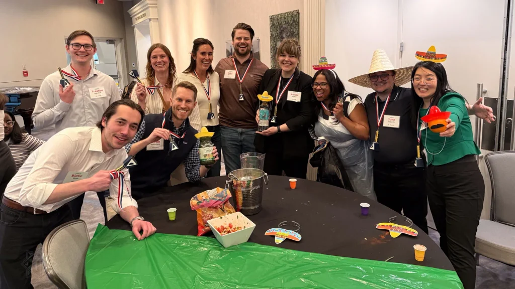 A smiling group wearing medals and festive hats poses with salsa jars around a table.