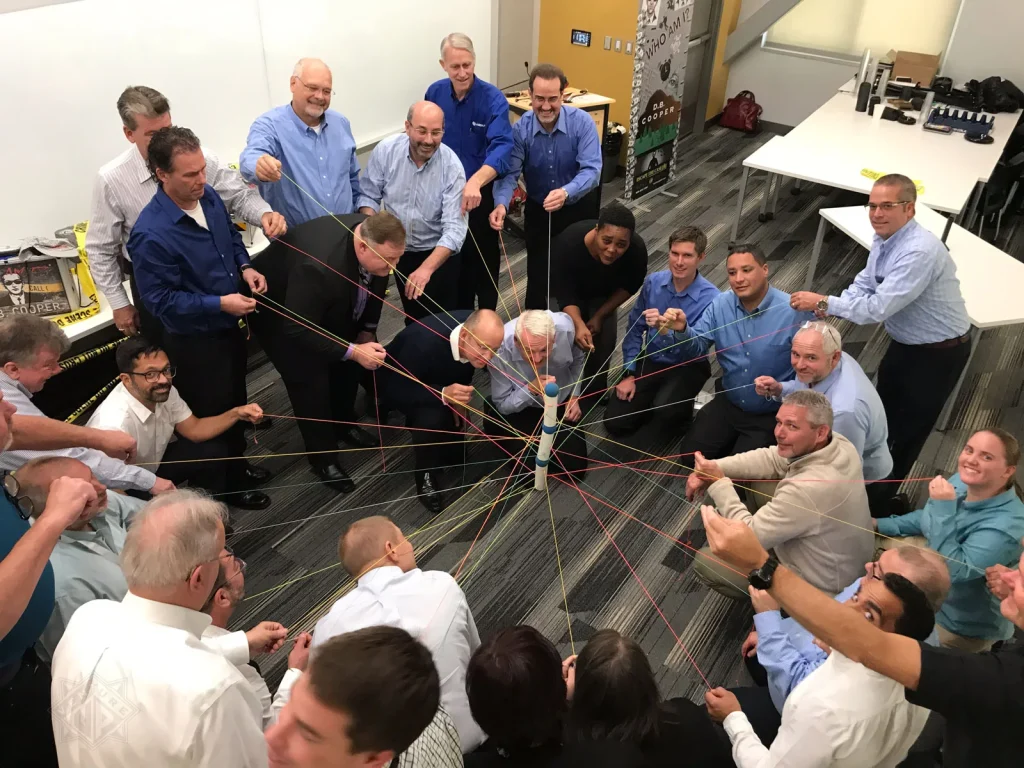 Group of people standing/kneeling in a circle, each holding colorful string attached to a central pole, creating a web-like network in a room.