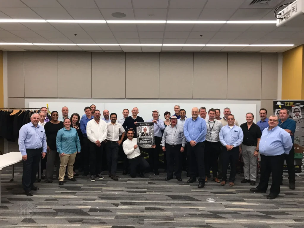 Group of coworkers posing for a team photo in a conference room, with one person kneeling center holding a poster plaque.