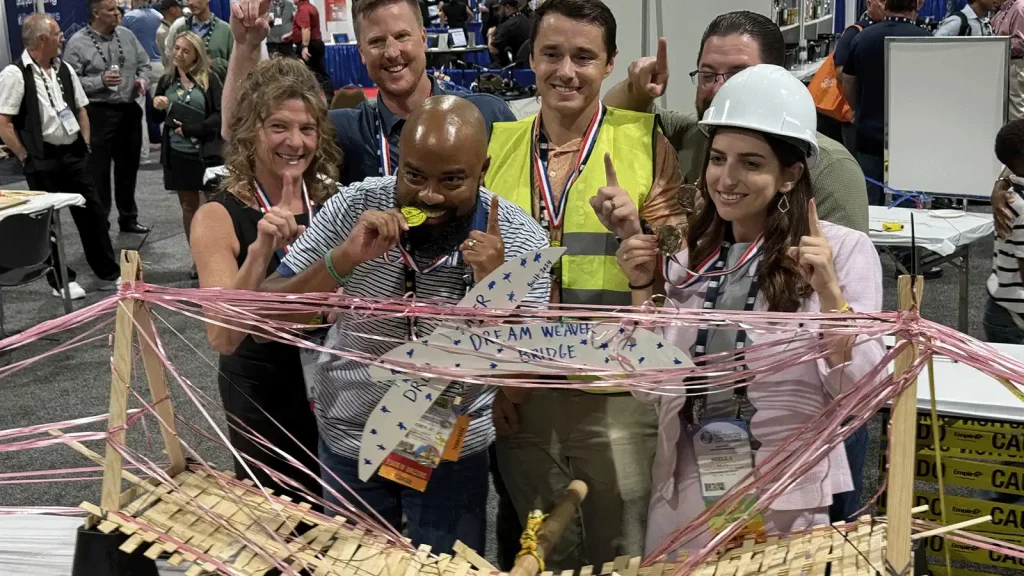 Group of diverse teammates posing with medals and raised index fingers, celebrating a project next to a pink string bridge model at a convention hall.