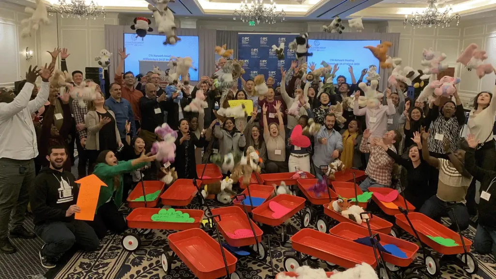 Large group of people at a conference throwing stuffed animals into the air, with red wagons filled with plush toys in the foreground.