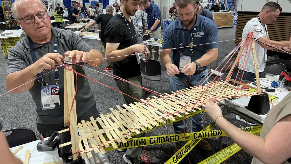 Group of people at a convention building a bridge from popsicle sticks and red string, with yellow caution tape around the workspace.