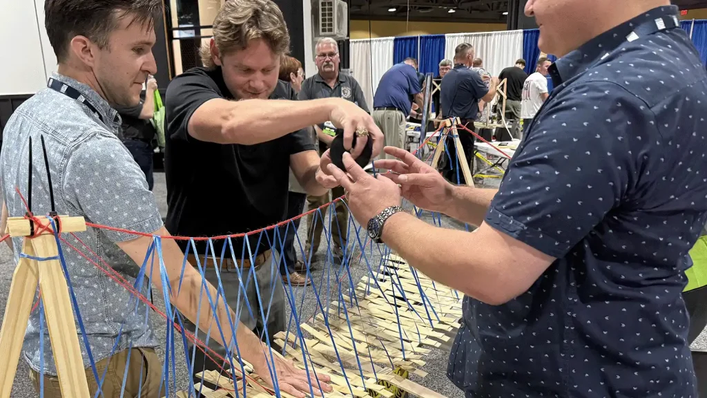 Three men tie blue and red strings to a wooden frame at a hands-on activity booth.