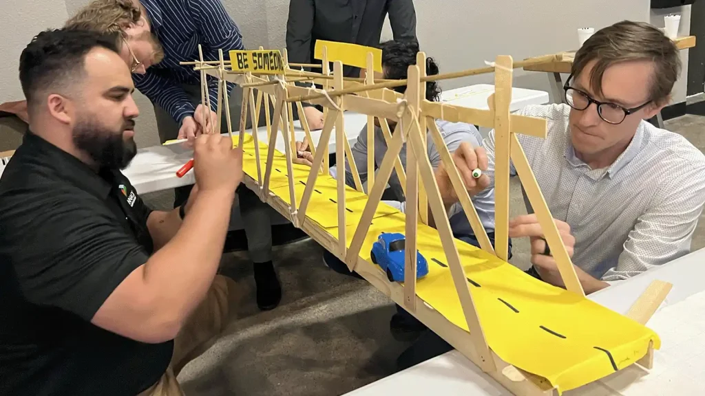 Team members build a long wooden bridge model with a bright yellow roadway and a small blue toy car on it, in a workshop setting. | Team Building Activities & Techniques