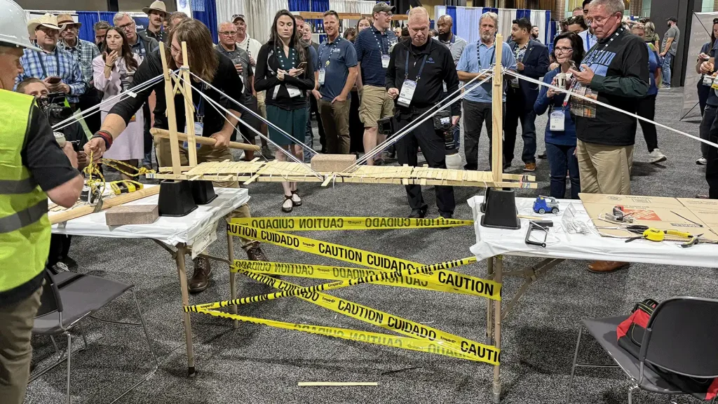 Crowd watches a wooden bridge-building demonstration at a convention, with yellow caution tape surrounding the setup on tables in a large room