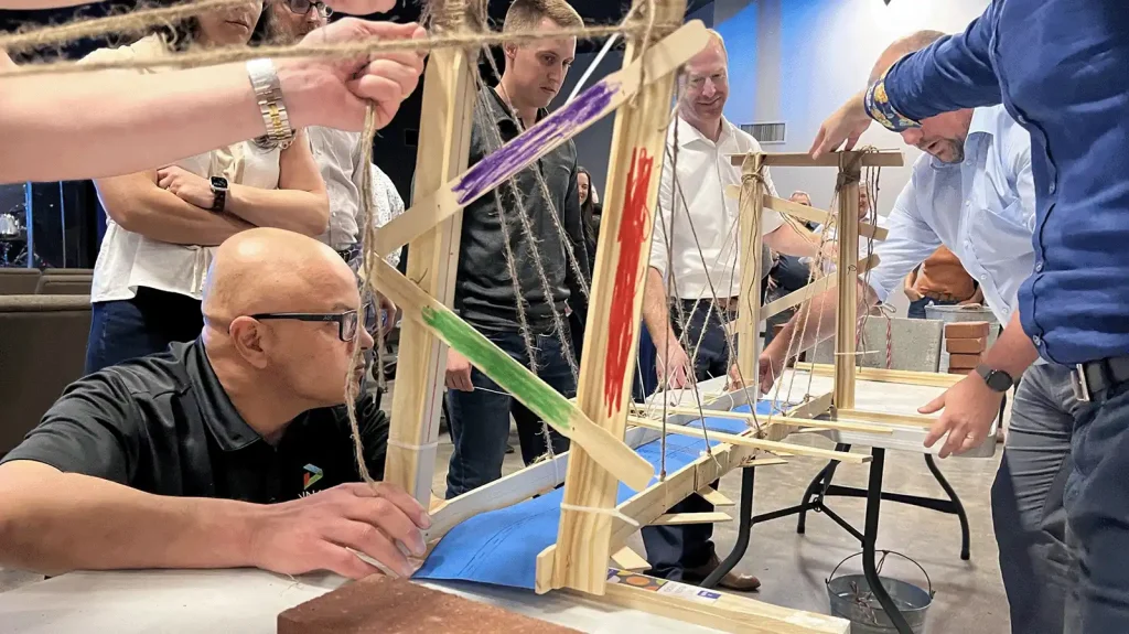 Group of adults watching a hands‑on weaving workshop with wooden loom frames and colorful strips of fabric or yarn nearby.