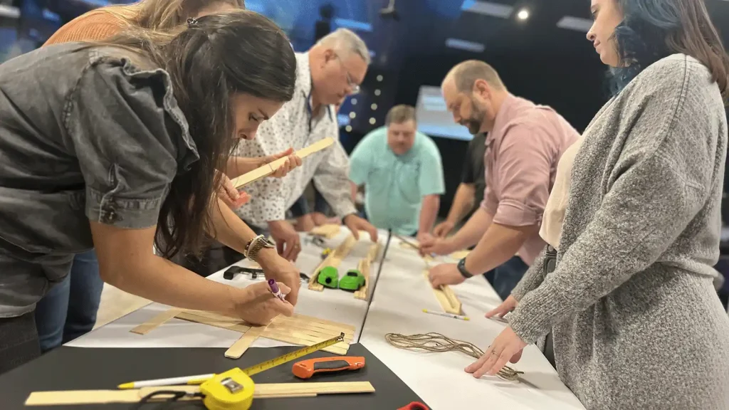 Group of adults gathered around a long table, crafting wooden pieces with rulers and tools in a workshop setting.