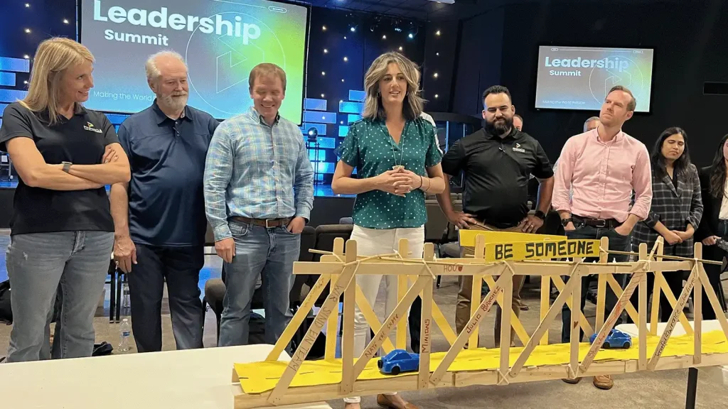 Group of people standing behind a wooden bridge model with toy cars, at a Leadership Summit backdrop labeled 'Be Someone' on the bridge.