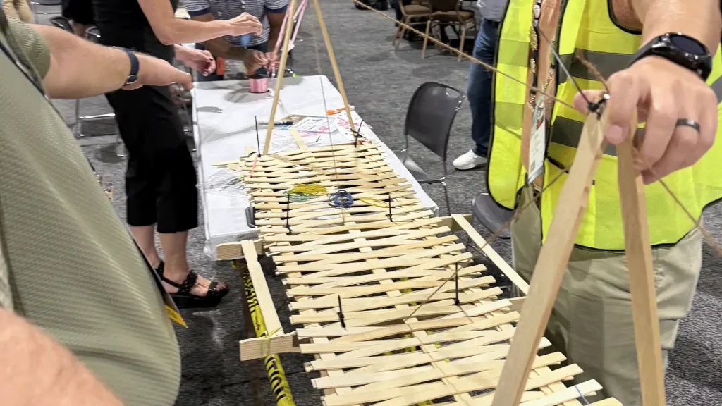 Group of people building a long popsicle-stick bridge connected with string on a table at a demonstration.