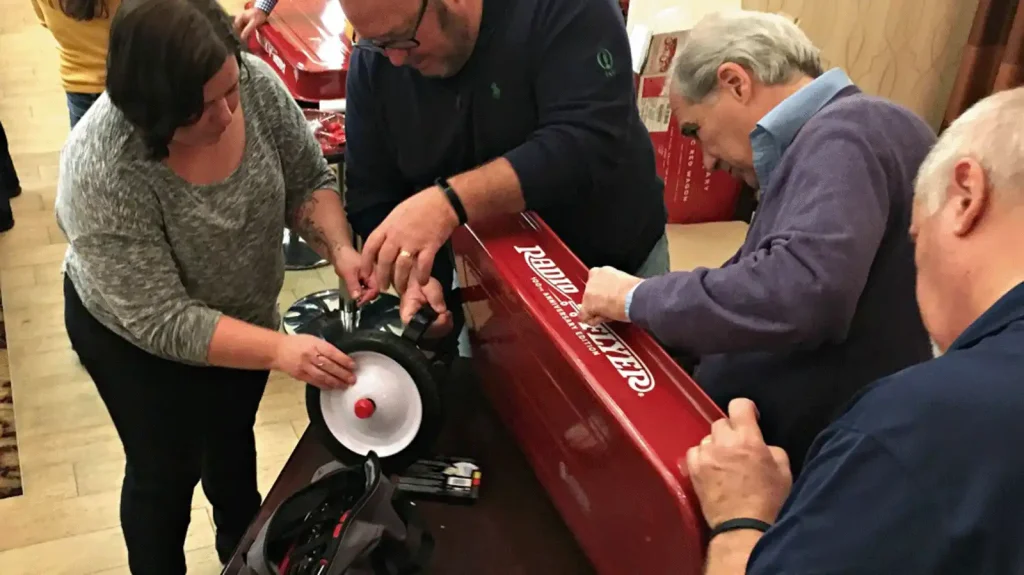 Group of adults gathered around a red boxed device, assembling it on a table; one person holds a wheel while others tighten parts.