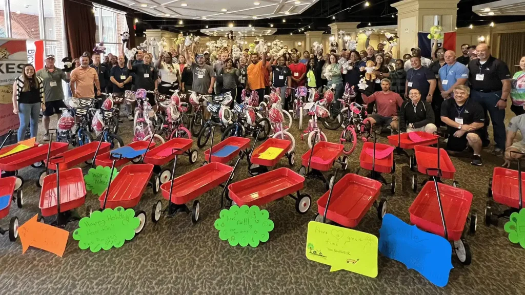Large group of volunteers and families posing behind a row of red wagons and small pink bikes in a banquet hall parking lot area (charity event).