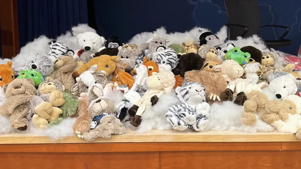 A large pile of stuffed animals (teddy bears and plush creatures) piled on a wooden table with white cotton-like fluff in the background.