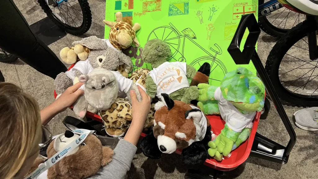 Child sorting through a red wagon full of stuffed animals, with a bright green bike-themed poster in the background behind them.