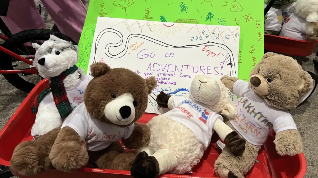 Group of stuffed bears in white T-shirts sitting in a red wagon, with a large handwritten poster reading 'Go on ADVENTURES!' in the background.