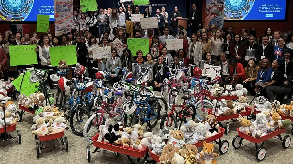 Group of volunteers posing with donated bikes and plush toys at a charity event, holding bright signs in the crowd behind them.