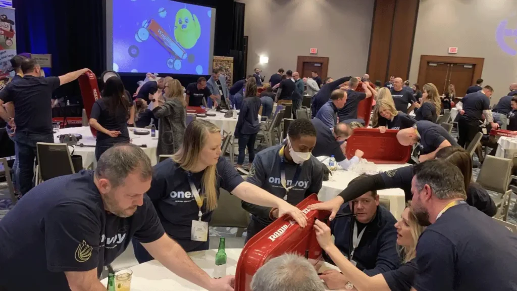 People at a large indoor event around tables, assembling a red plastic object together as part of a team activity; stage and screen in the background.