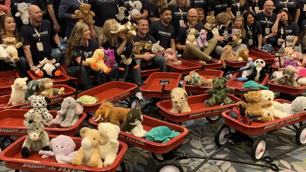 Group of volunteers in matching shirts posing with red wagons filled with stuffed animals for a donation drive.