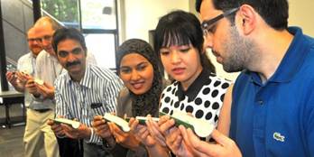 Five adults of diverse backgrounds standing side by side in a casual setting, each looking at a small rectangular device (likely smartphones) in their hands. | Team Building Activities & Techniques