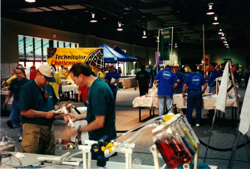 Crowded convention hall with volunteers in blue shirts at science fair booths; a yellow banner reads 'Technicolor Challenge' in the background. | Team Building Activities & Techniques
