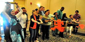 Group of coworkers in a conference room holding a long orange inflatable object as part of a team-building activity, smiling and engaged in the exercise. | Team Building Activities & Techniques