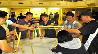 Group of people gathered around a table constructing a yellow wooden framework together in a workshop setting. | Team Building Activities & Techniques