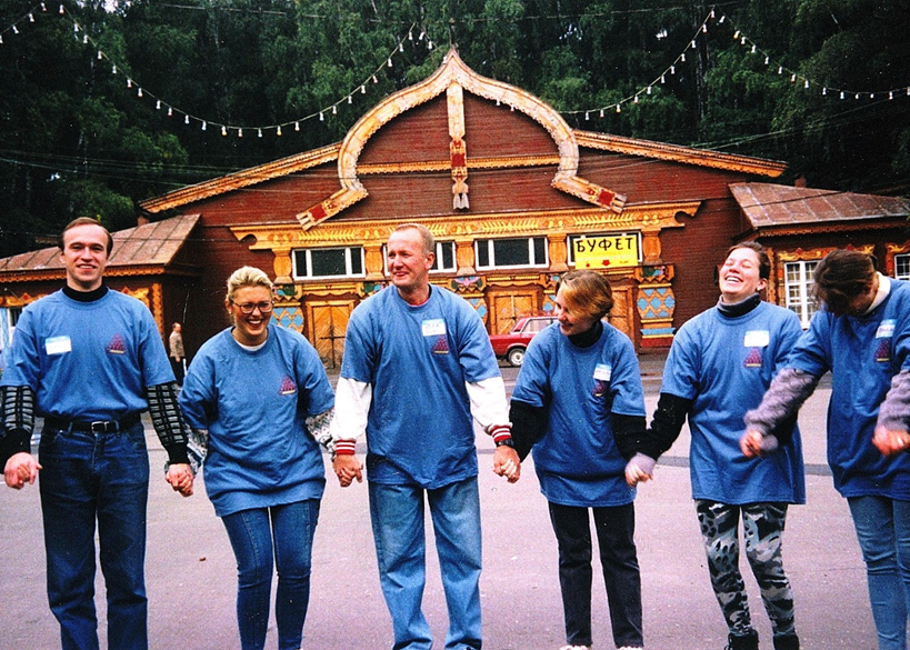 Six adults in matching blue shirts form a circle, holding hands in front of a wooden, festively decorated building with string lights. | Team Building Activities & Techniques